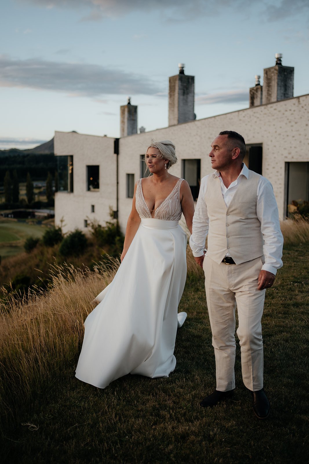 A couple holding hands, dressed formally, walking on grass with a modern building and hills in the background.