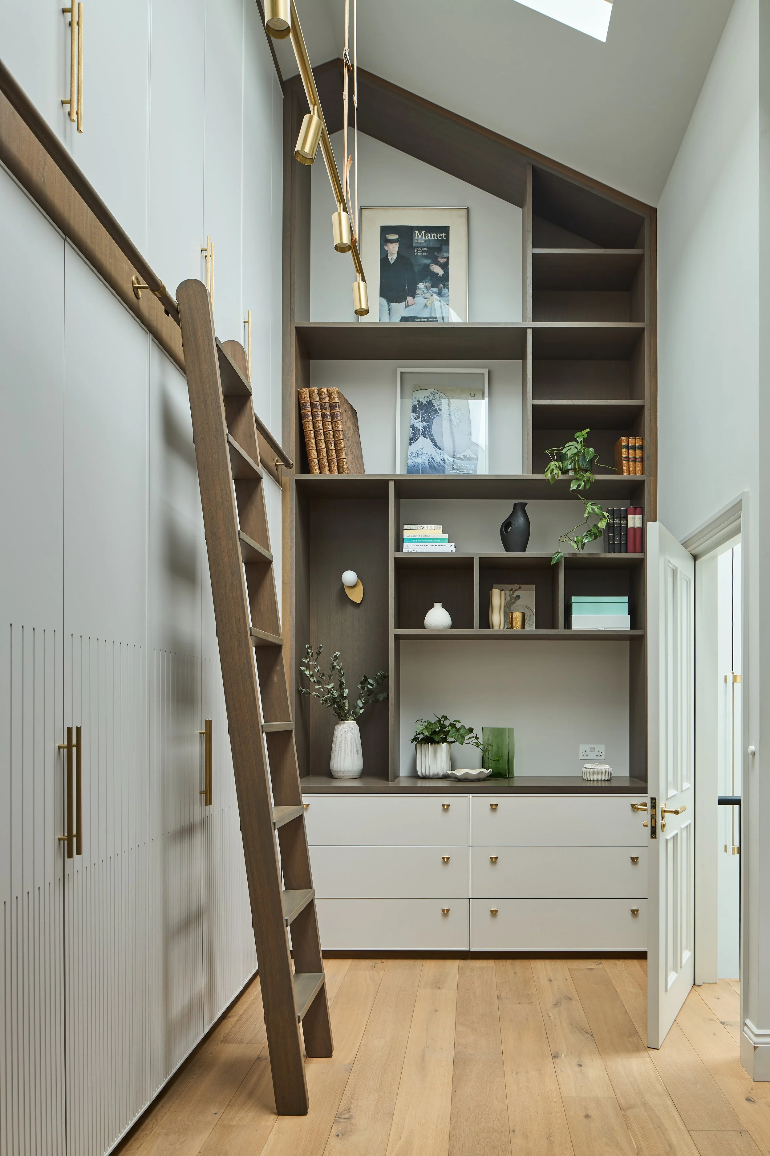 Wall of bespoke joinery with asymmetrical shelving in contrasting finishes, brass handles and a library ladder.