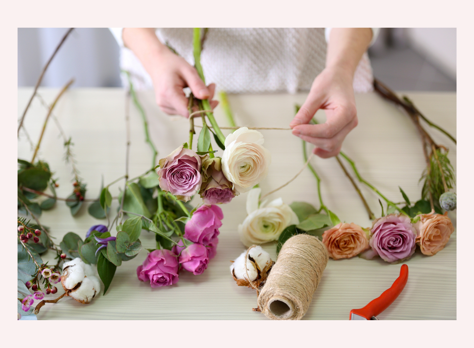 Person arranging a floral bouquet with roses and cotton stems on a light-colored table.
