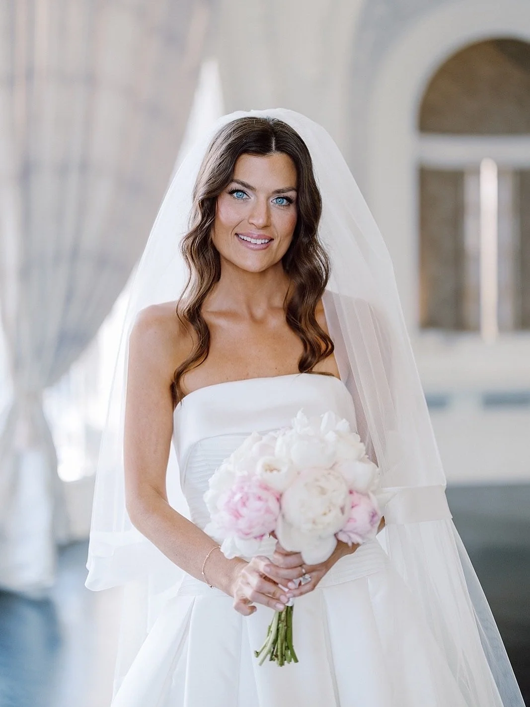 A smiling bride with blue eyes and wavy brown hair in a strapless white wedding dress holding a bouquet of white and pink peonies inside a bright, elegant venue.