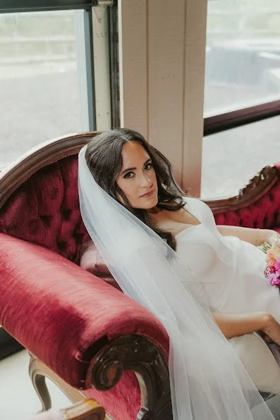 Bride lying on a vintage red velvet sofa with a white veil, holding a bouquet of pink flowers, near large windows.