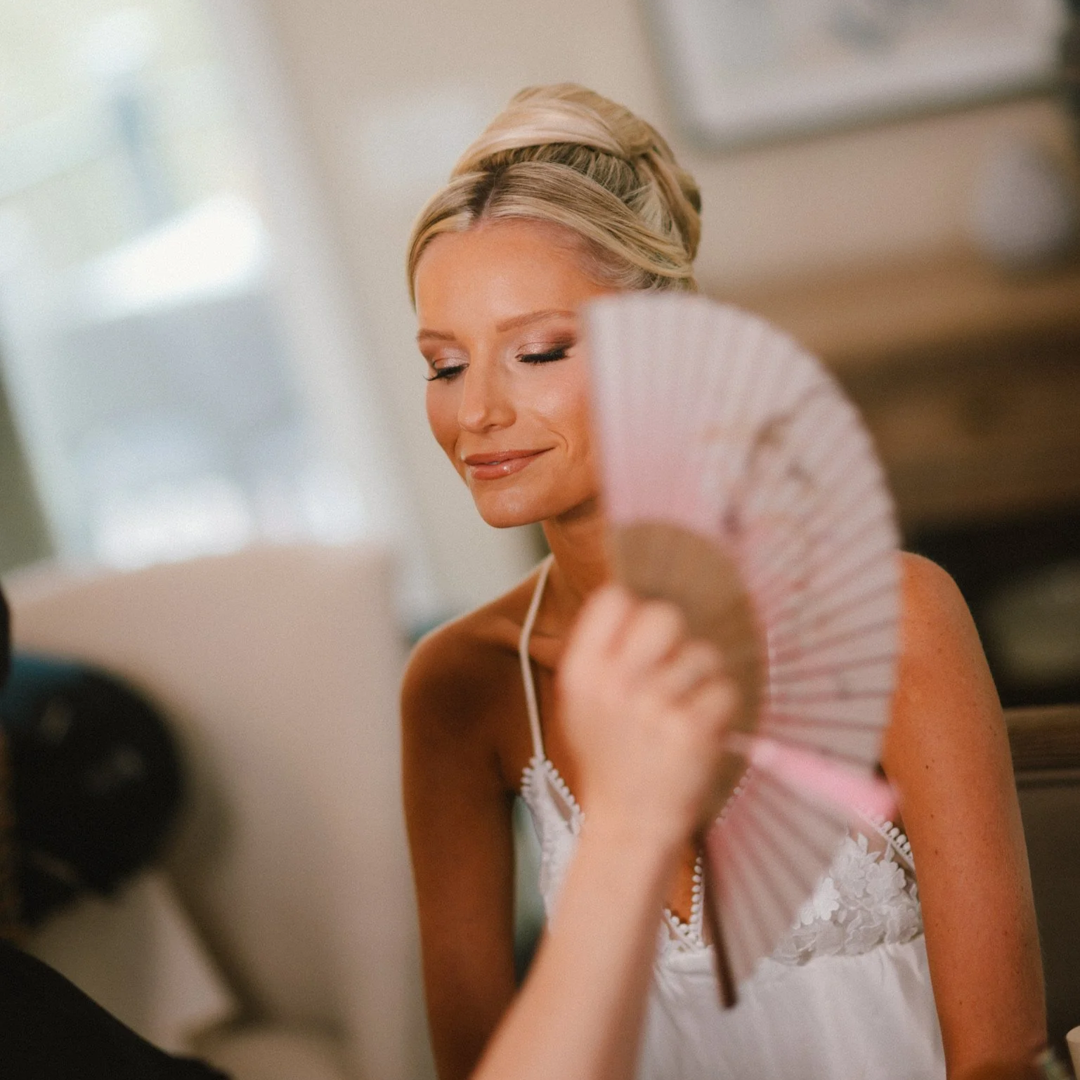 A blonde woman with a her hair styled in an elegant updo, wearing a white dress, holding a pink fan, with eyes closed and a gentle smile.