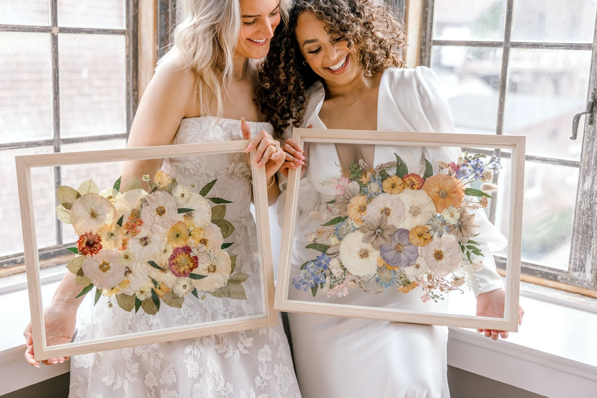 Two women in white dresses holding framed floral arrangements, smiling and leaning close together, standing near large windows.