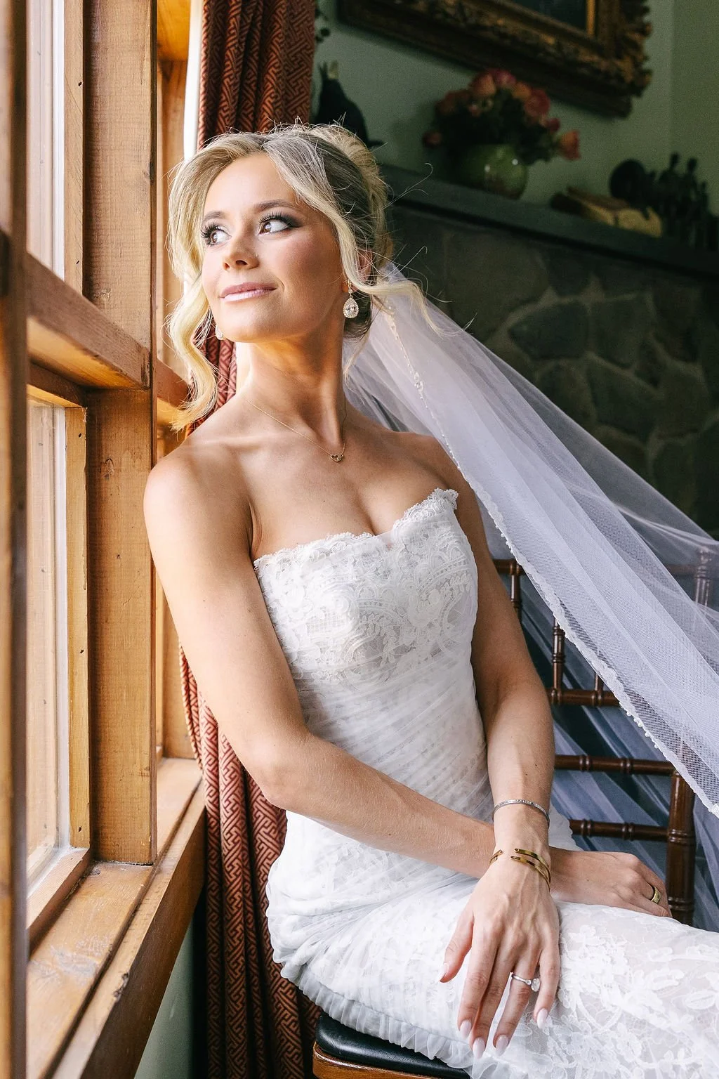 A bride sitting by a window in a white wedding dress, gazing outside.