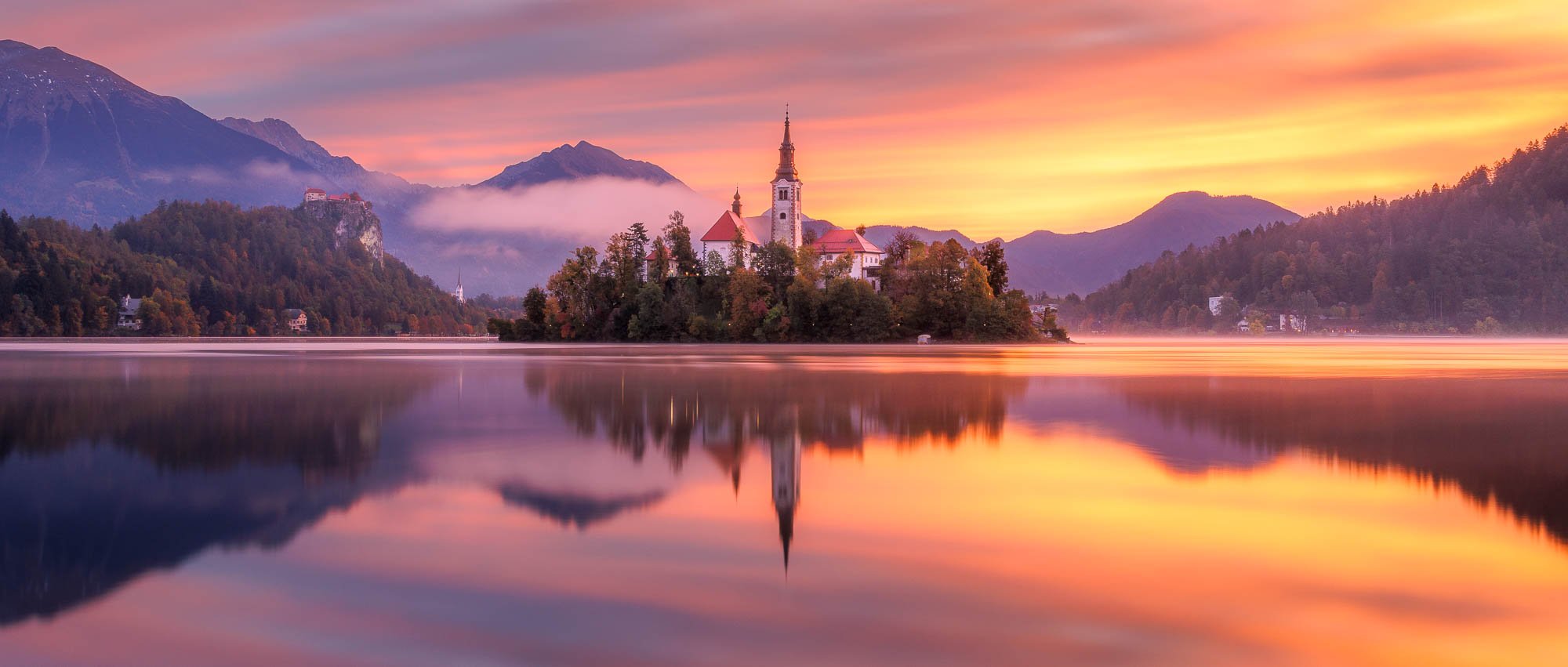 Lake Bled island with church at sunrise, surrounded by mountains and autumn foliage, reflecting on the water's surface.