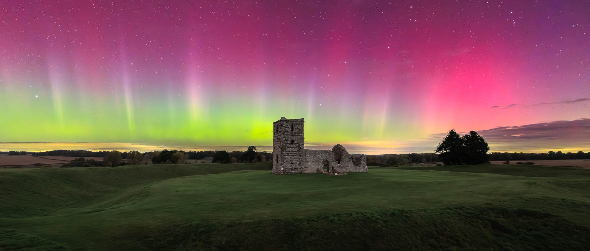 Ruined stone building on grassy field under vibrant aurora with pink and green lights.