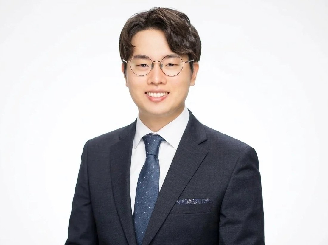 A young man with dark hair, glasses, and a bright smile dressed in a dark suit, white shirt, and navy tie with small white and teal dots, standing against a blue background.