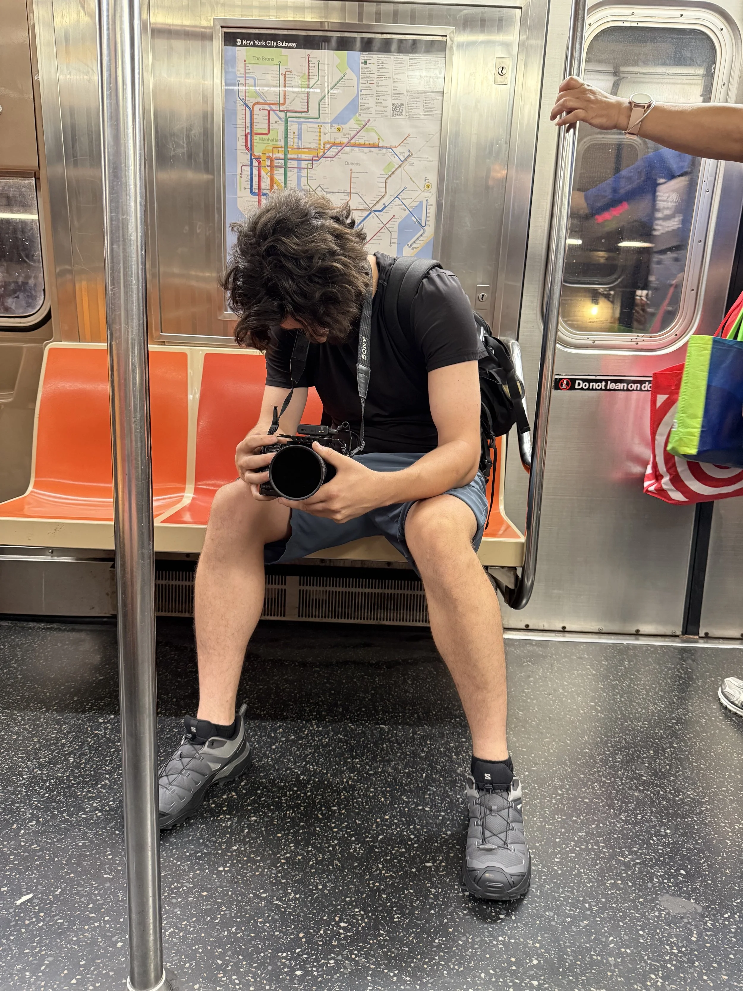 A person sitting on a subway train seat looking at a camera, with a NYC subway map behind them.