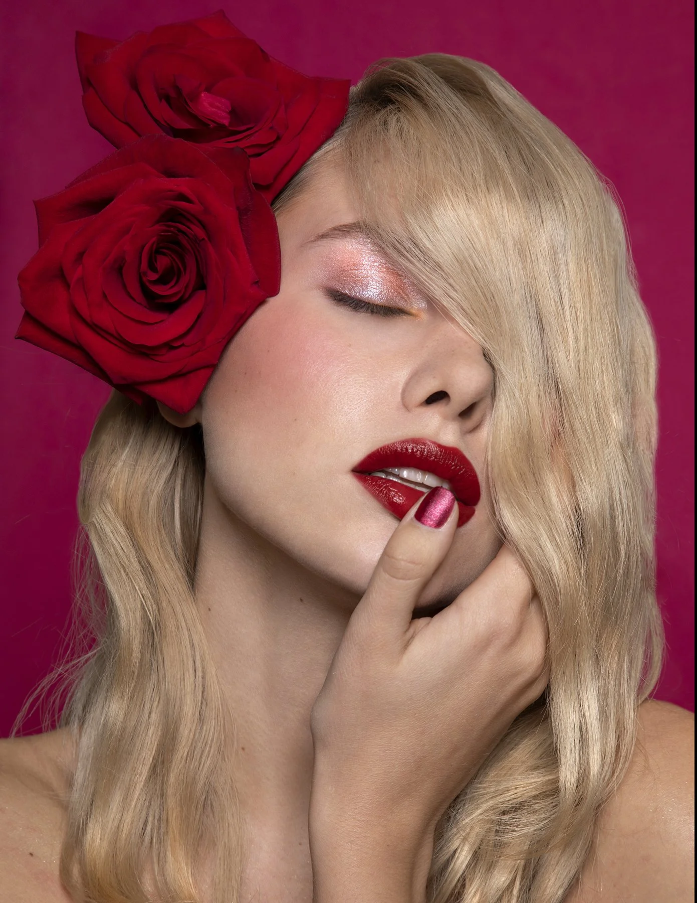 A woman with long blonde hair and red lipstick, near her lips, is touching her face. She has makeup with pink eyeshadow, and two large red roses are tucked in her ear. The background is pink.