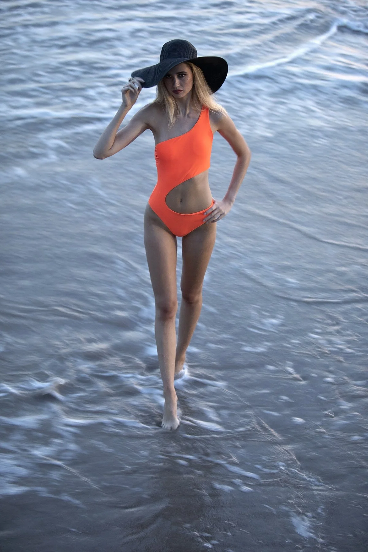 A woman in an orange swimsuit and a large black sun hat standing in shallow ocean water.