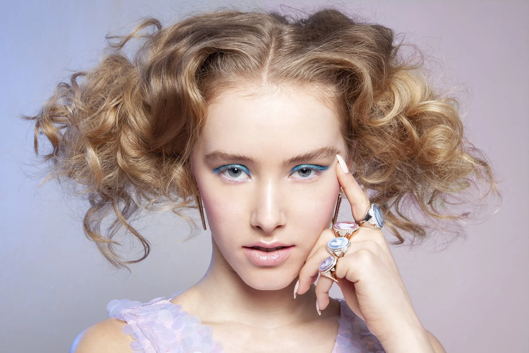 A young woman with curly blonde hair wearing blue eyeliner and several colorful rings, looking directly at the camera with her hand touching her face.