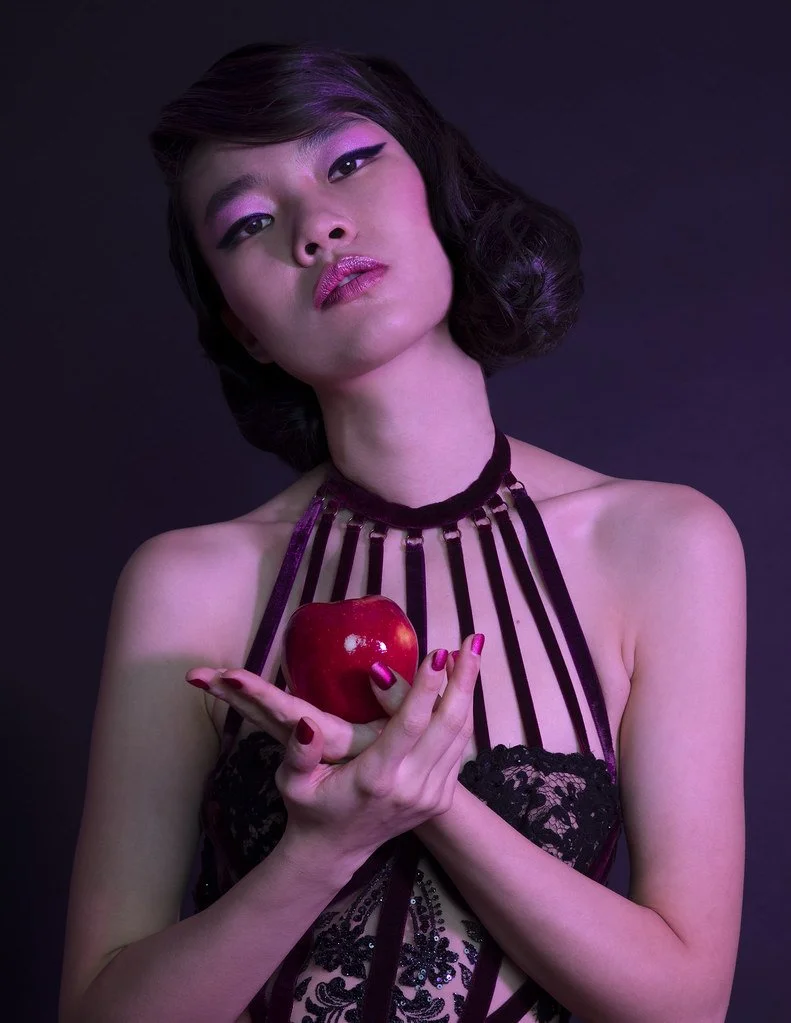 A woman with short wavy hair, makeup, and pink lipstick, holding a red apple in her hands, against a dark background.