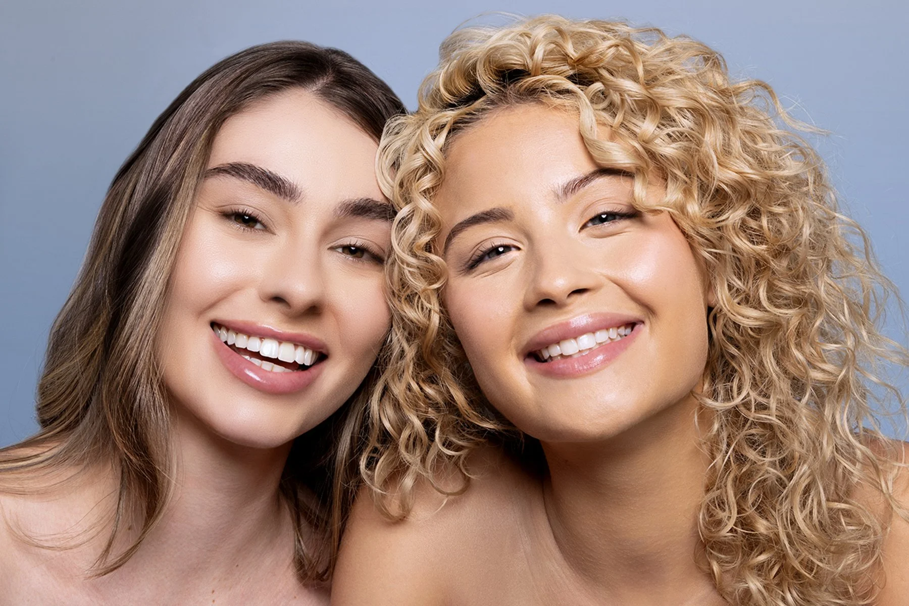 Close-up of two smiling women with clear skin, one with straight brown hair and the other with curly blonde hair, posing together against a light blue background.