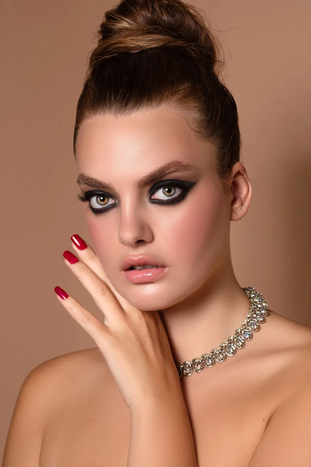 Close-up portrait of a woman with styled hair, bold eye makeup, and a sparkling necklace, posing with her hand near her face against a neutral background.
