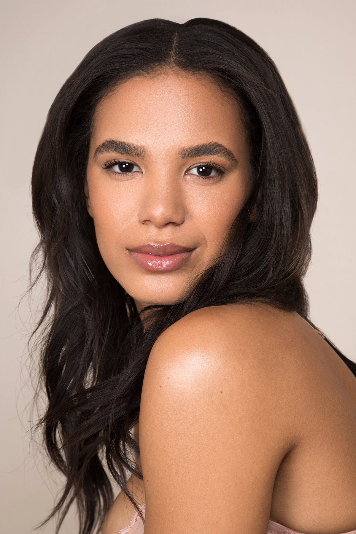 Close-up of a woman with long dark wavy hair, light skin, full lips with gloss, and dark eyes. She is looking slightly over her shoulder, with bare shoulders and a neutral background.