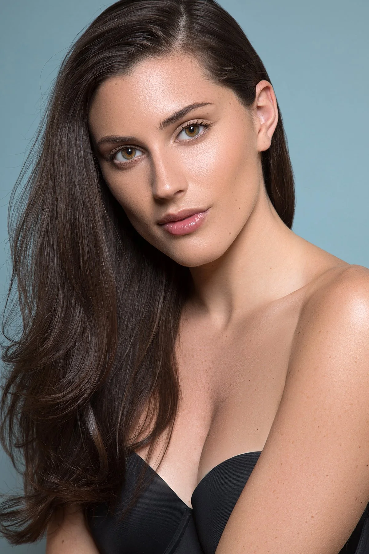 A young woman with long, dark brown hair and light skin, wearing a black strapless top, posing against a plain light blue background.