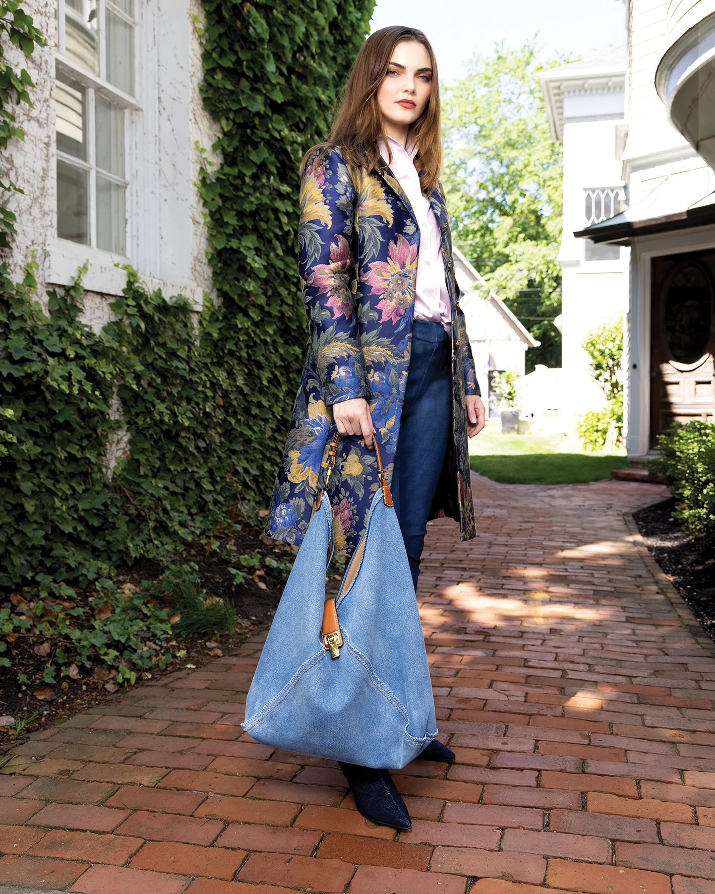 A woman standing on a brick sidewalk in front of a house with ivy-covered walls. She is wearing a floral-patterned coat, jeans, and dark boots, and holding a large denim tote bag.