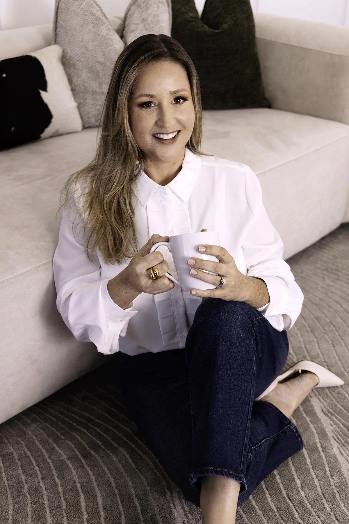 A woman sitting on the floor next to a beige sofa, smiling, holding a white mug, wearing a white blouse and blue jeans.
