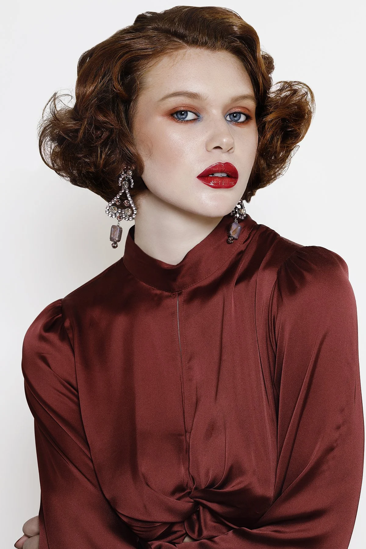 A woman with curly red hair and blue eyes wearing red lipstick, large earrings, and a satin red dress against a plain white background.