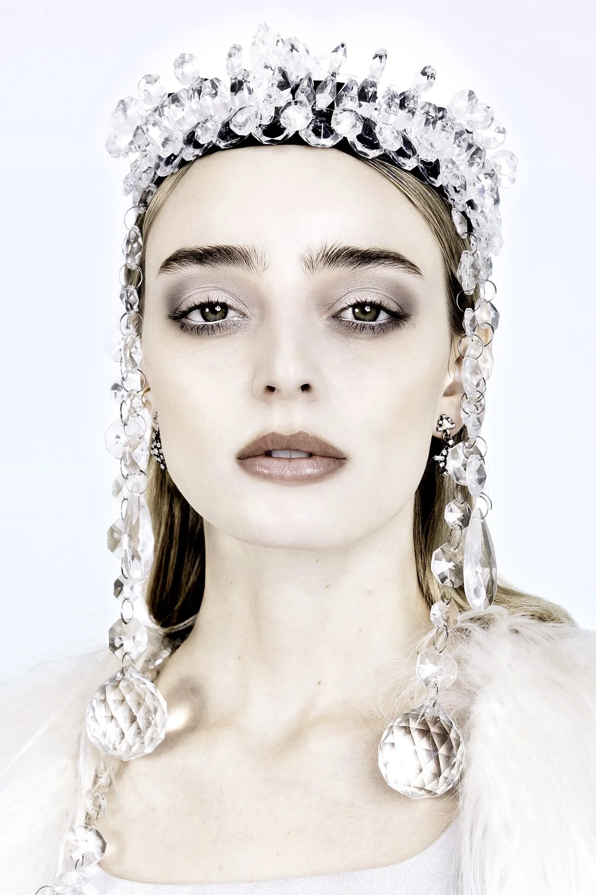 A woman wearing a crown and earrings made of large clear crystals, with a soft makeup look and light-colored clothing.