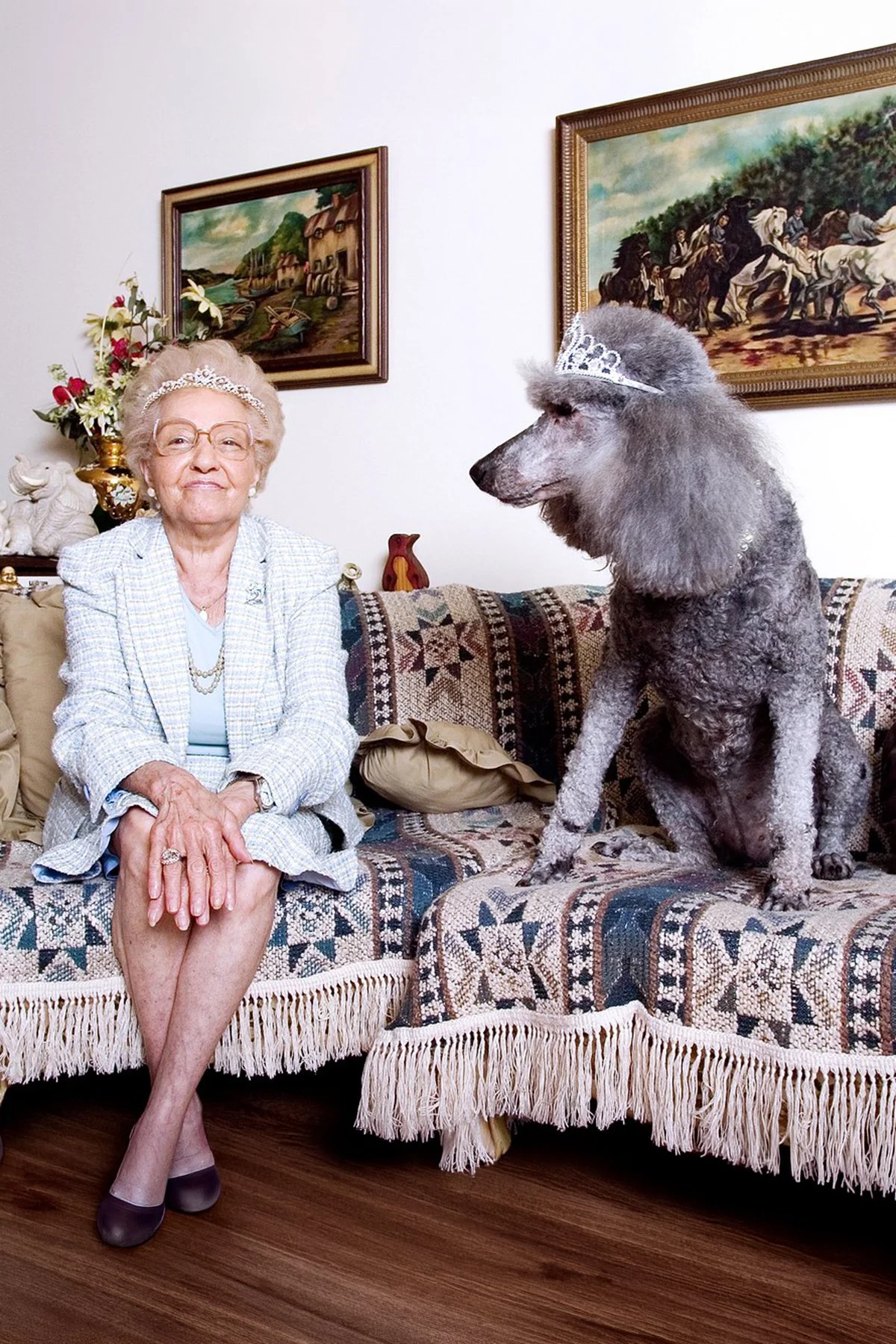 An elderly woman wearing a tiara and glasses, seated on a patterned sofa with her legs crossed. Next to her is a large dog wearing a tiara, sitting on the sofa. The background features two framed landscape paintings on the wall and decorative items o
