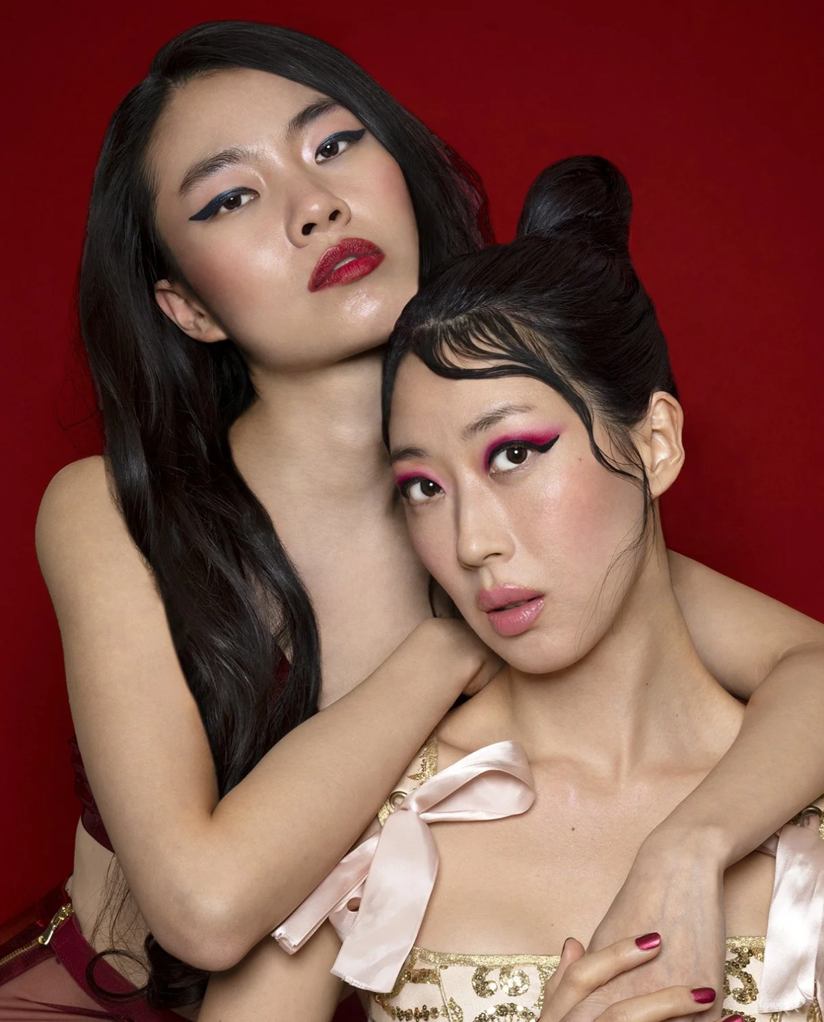 Two women with bold makeup, one with dark hair and the other with black hair styled in buns, posing closely with a red background.