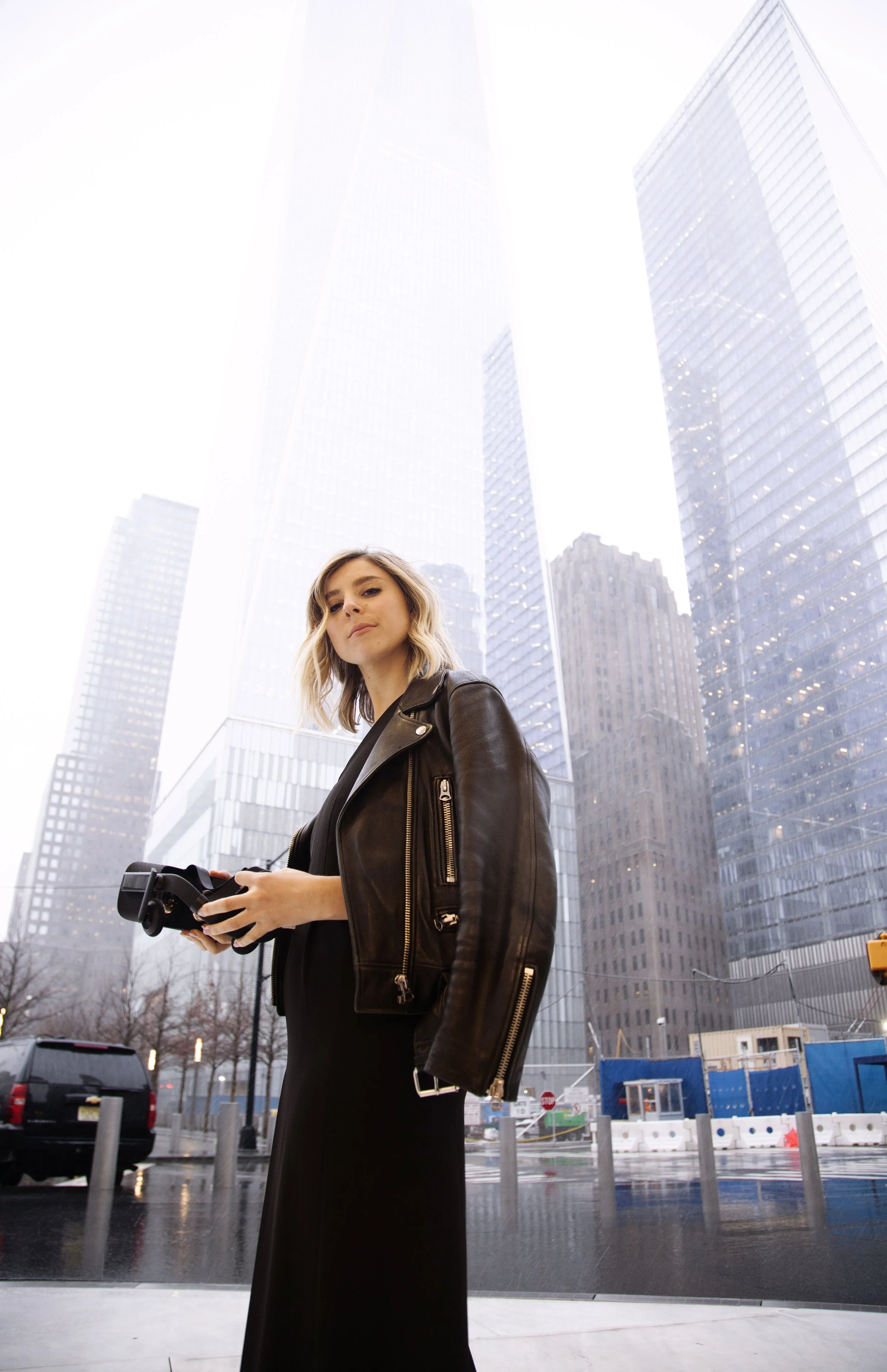 A young woman with blonde hair wearing a black leather jacket holding a camera or drone remote control, standing outside in an urban area with tall glass skyscrapers and construction site.