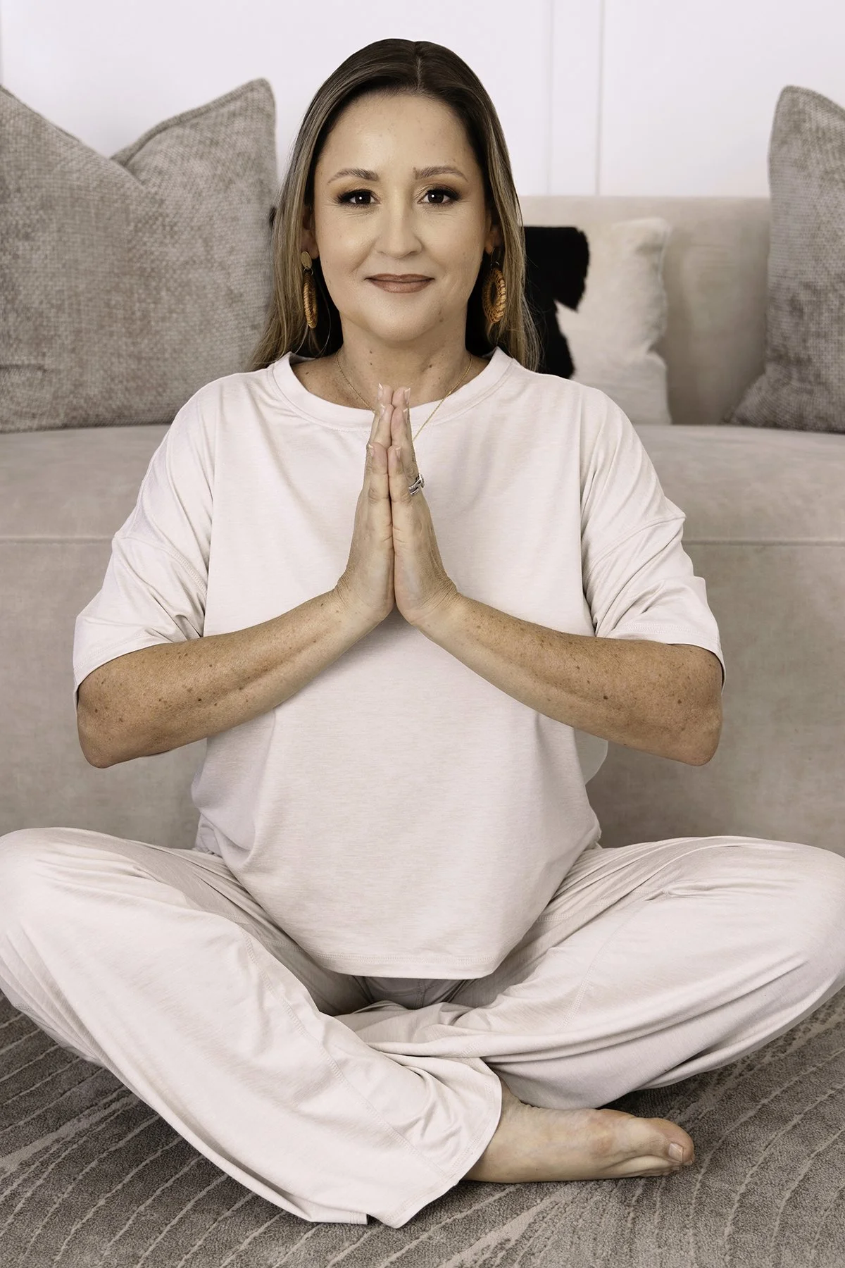 A woman practicing yoga in a seated cross-legged position on a carpeted floor with her hands pressed together in a prayer pose, smiling gently and looking straight ahead in a neutral indoor setting.