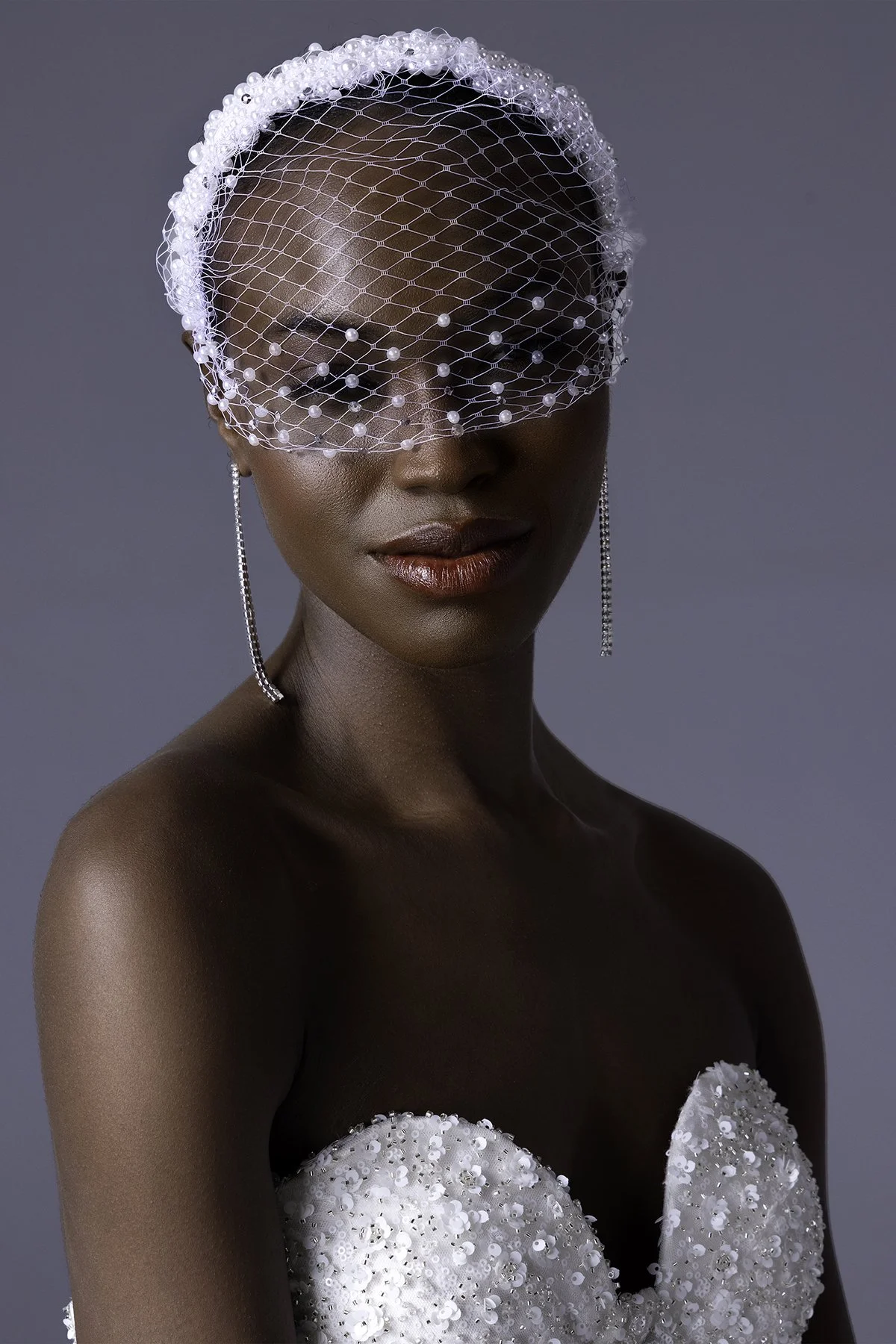 A woman in a white embellished dress with sequins and beads, wearing a white netted fascinator with pearls, long dangling earrings, and red lipstick.