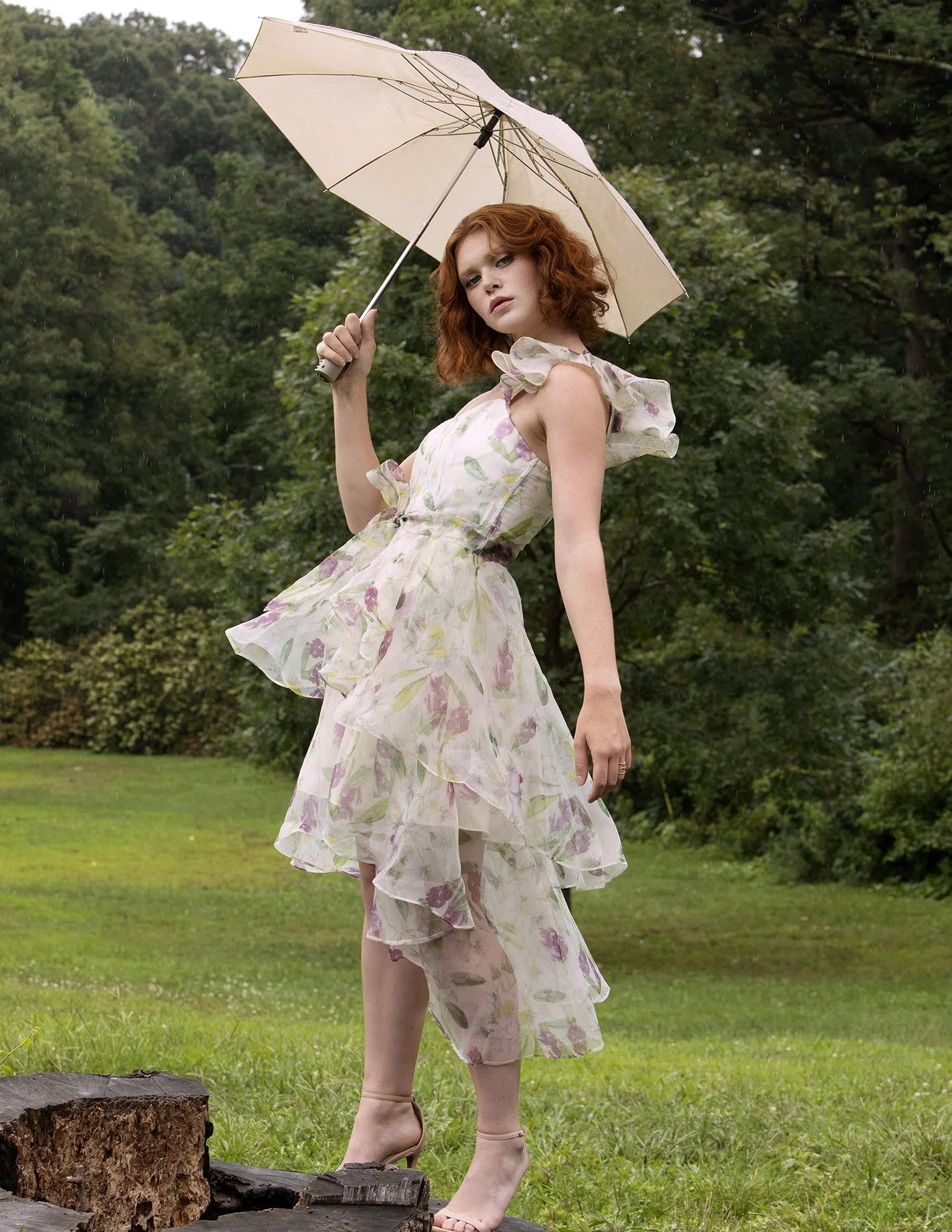 A woman with red curly hair standing outdoors on grass, holding a beige umbrella, wearing a light flower-patterned dress with ruffles and sheer fabric, and beige high-heeled shoes, with greenery and trees in the background.