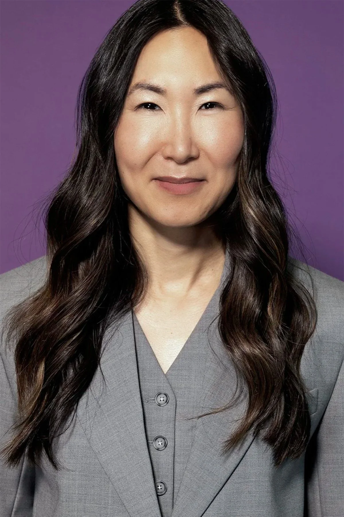 Head and shoulders portrait of an Asian woman with long wavy dark hair, wearing a gray button-up top, set against a purple background.