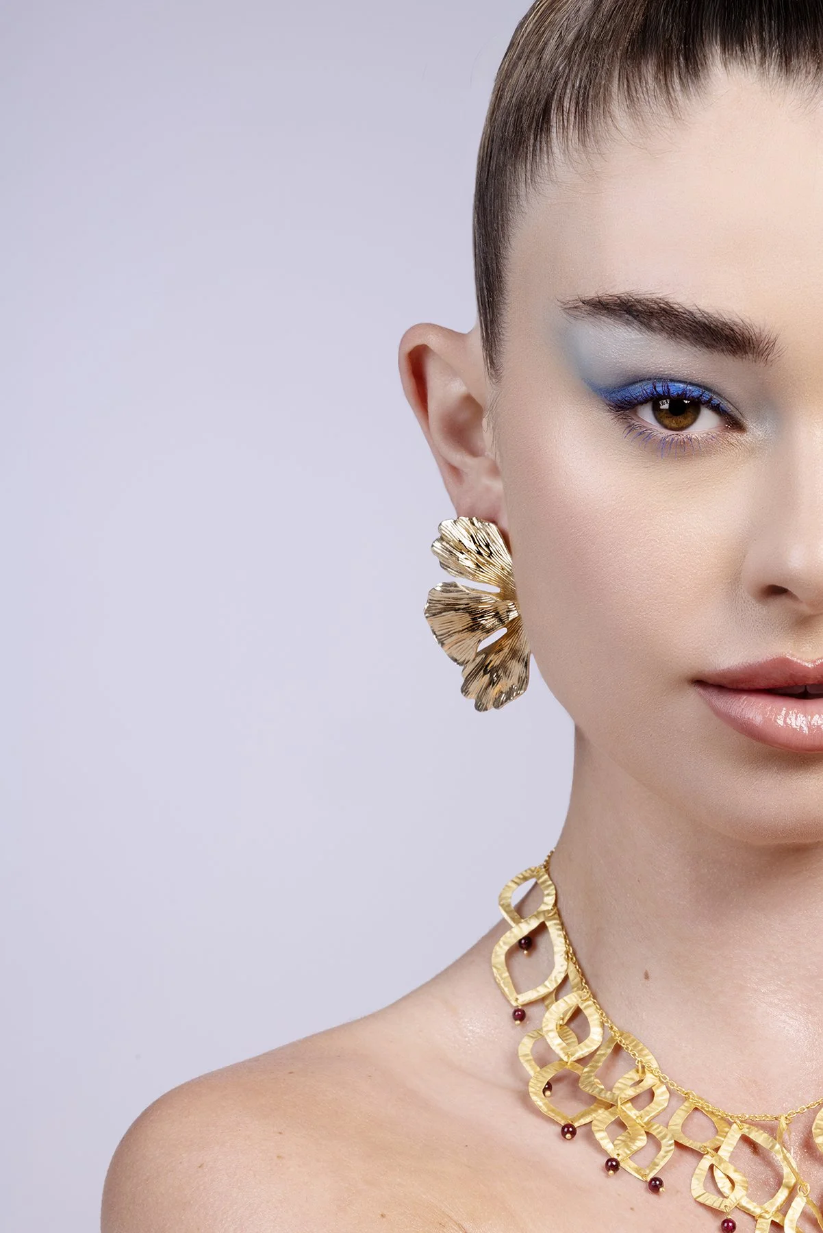 Close-up of a woman wearing gold jewelry, with a gold leaf-shaped earring, gold necklace with linked designs, and makeup featuring blue eyeshadow.