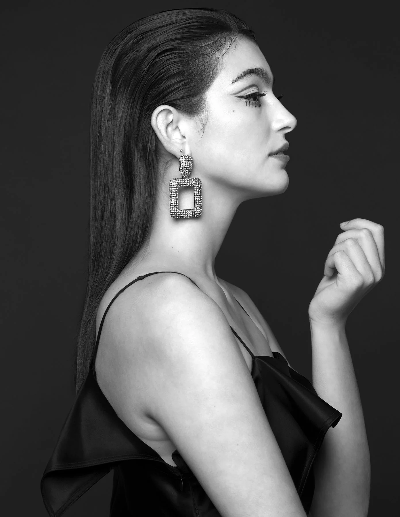 Profile of a woman with dark hair, wearing large square earrings adorned with rhinestones, dressed in a black satin top with thin straps, against a dark background, in black and white.