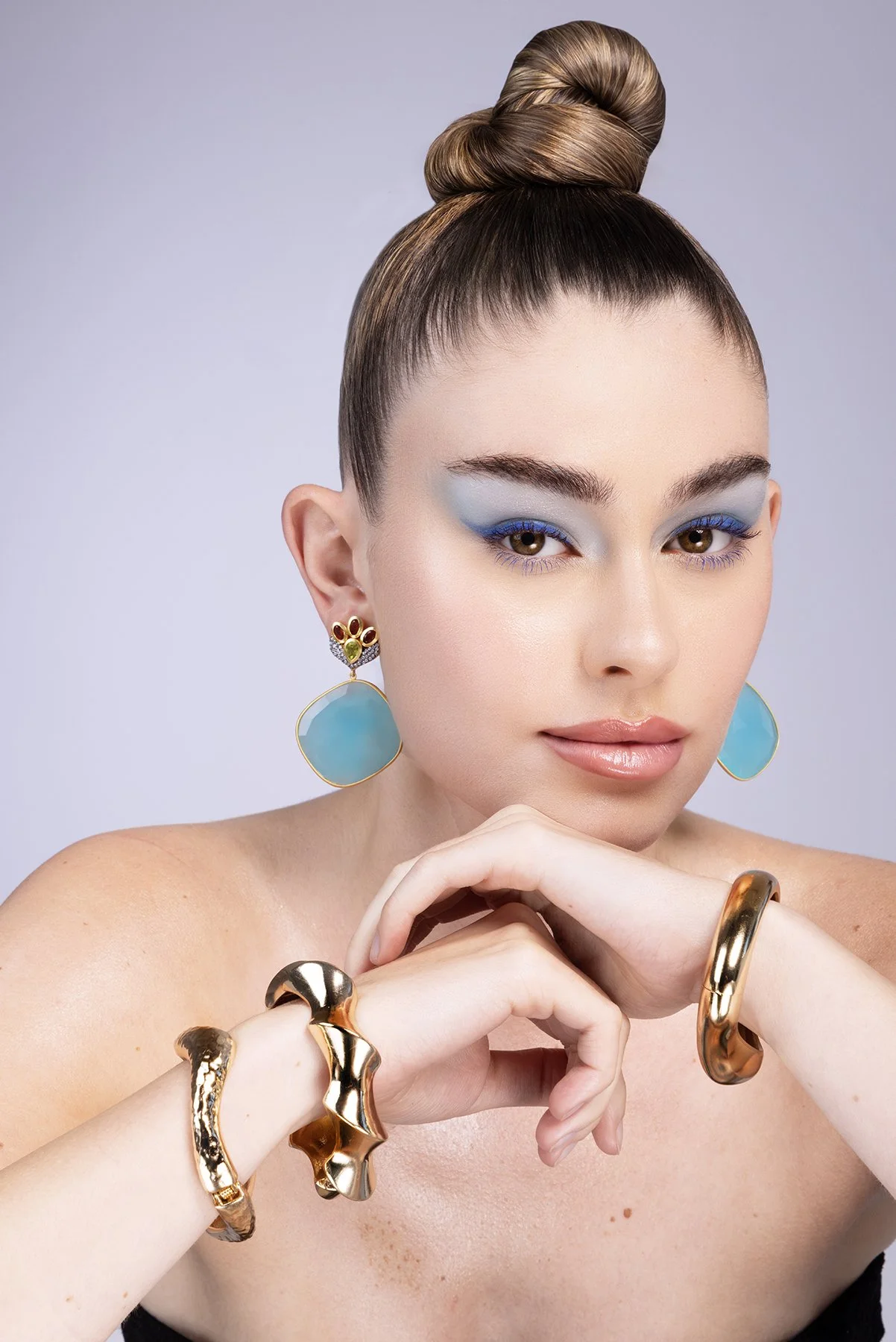 A woman with styled brown hair in an updo, wearing bold blue eye makeup and large turquoise earrings, with gold jewelry on her hands and wrists, and posing with her hands near her face.