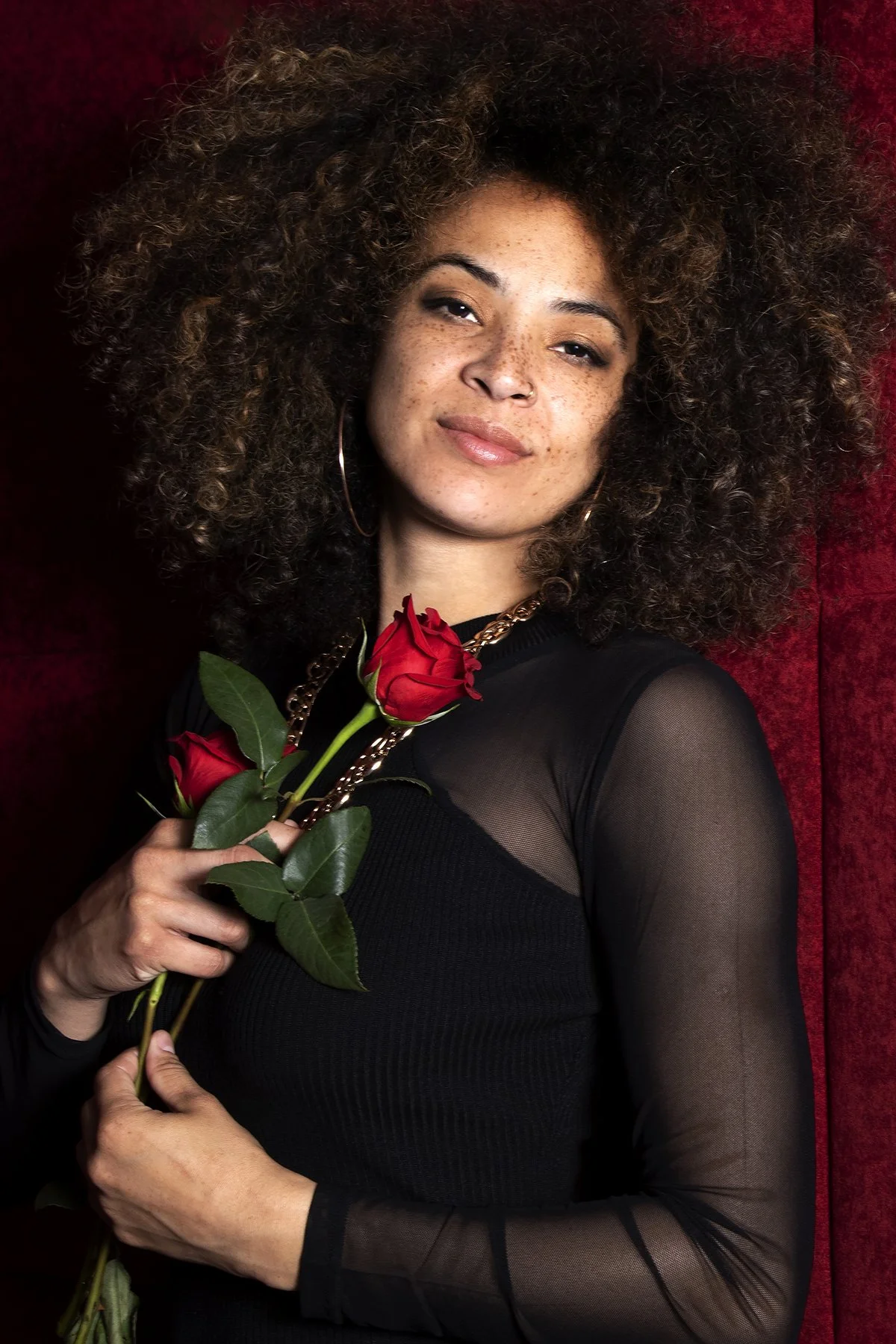 A woman with curly hair holding three red roses, wearing a black top with mesh sleeves, standing against a red velvet background.