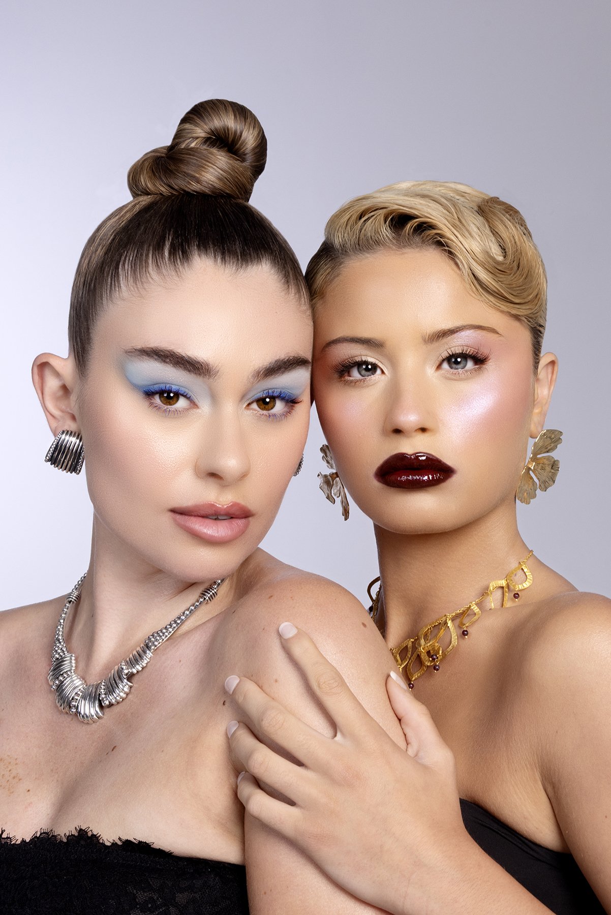 Two women with styled hair and elaborate jewelry posing closely together. One has makeup with blue eyeshadow, and the other has dark red lipstick and golden earrings.