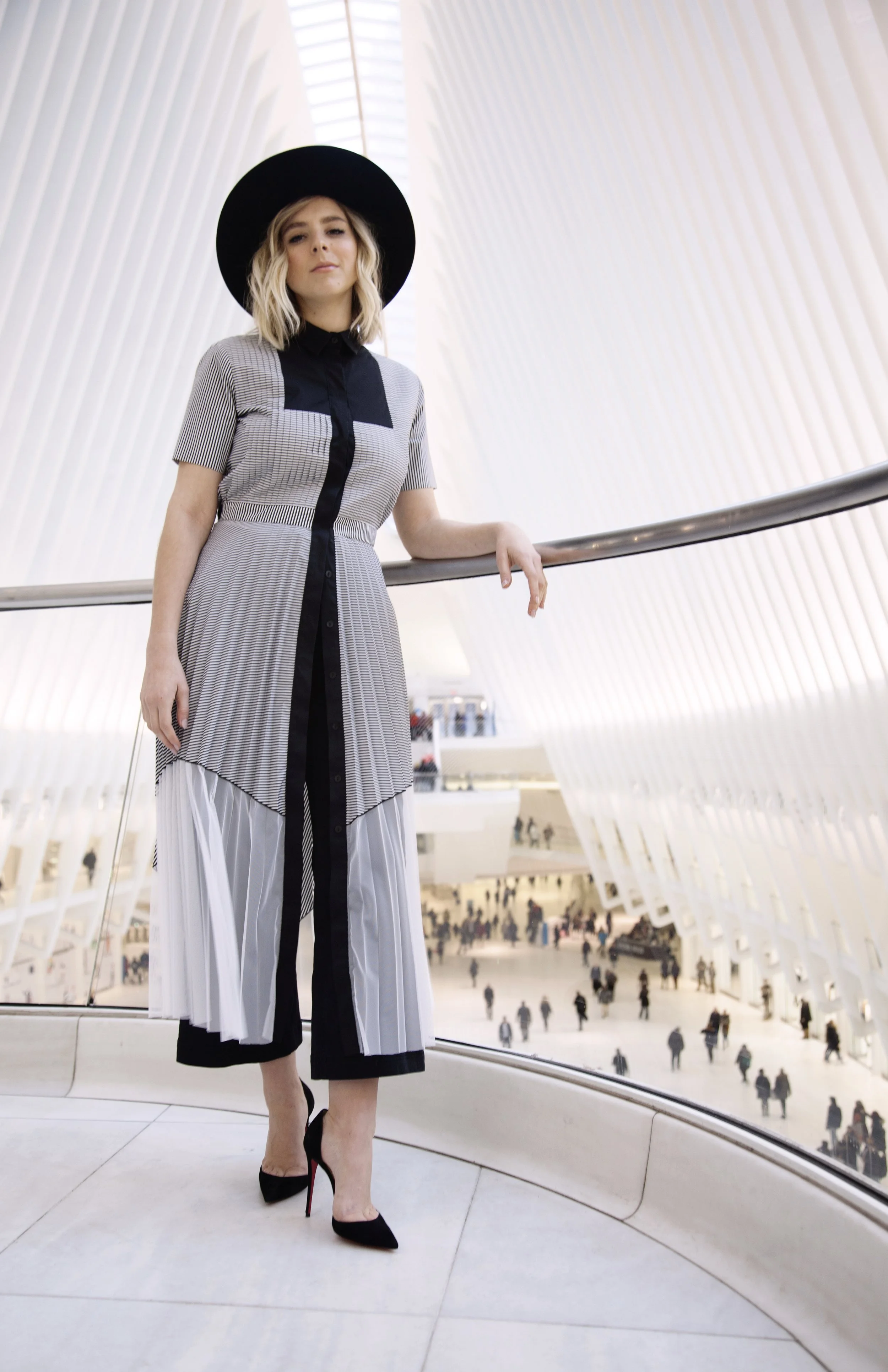 A woman wearing a black wide-brim hat, a black and gray striped dress, and black high heels with red soles standing on a curved balcony inside a modern building with a large glass roof and a busy crowd below.