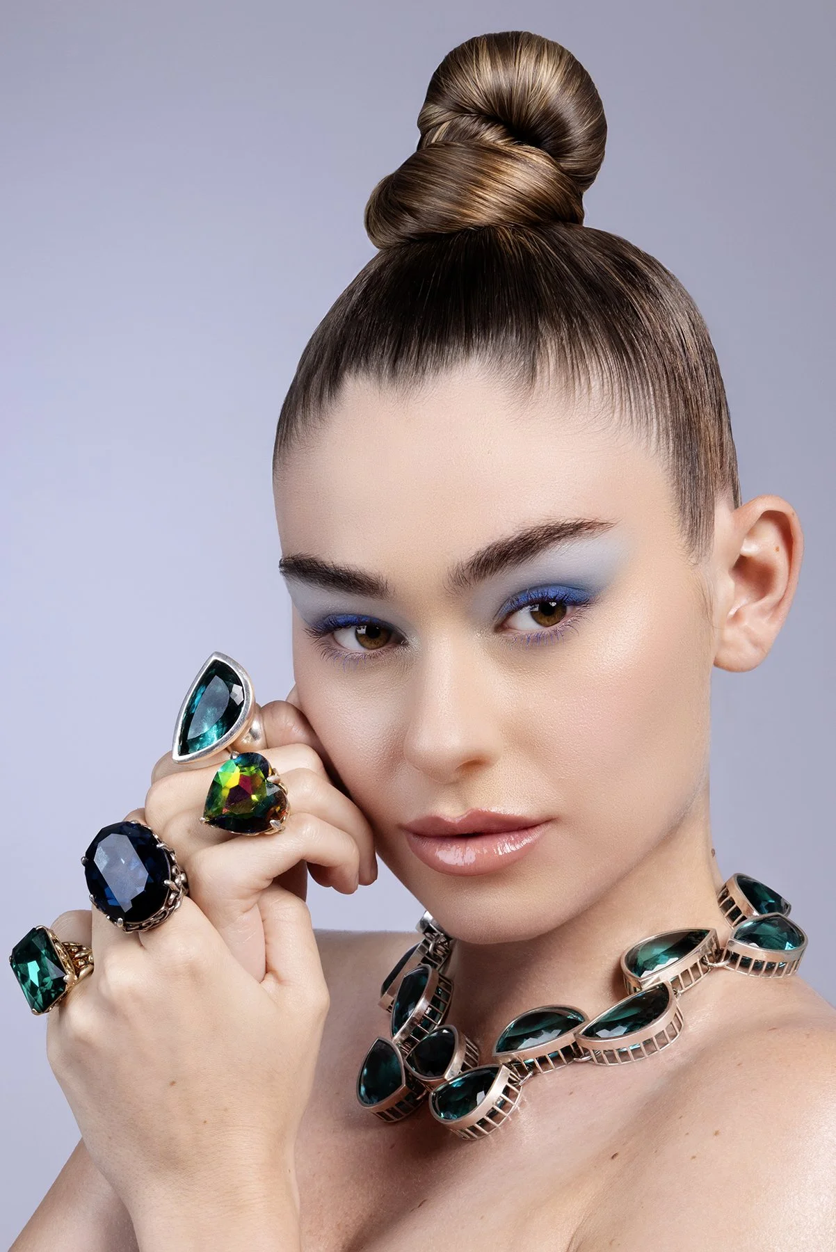 A woman with brown hair styled in a high bun, wearing bold blue eyeshadow and glossy lips, displays multiple rings and a necklace with large green gemstones, holding her hand near her face.