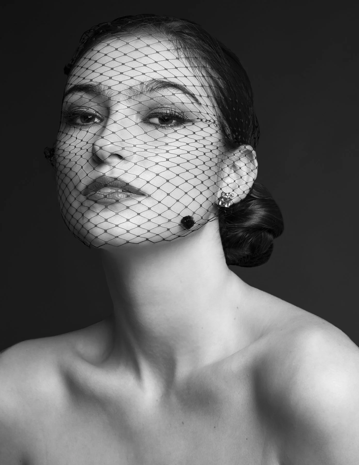 Black and white portrait of a woman with a decorative veil covering her face, wearing earrings, and her hair styled in an elegant updo.