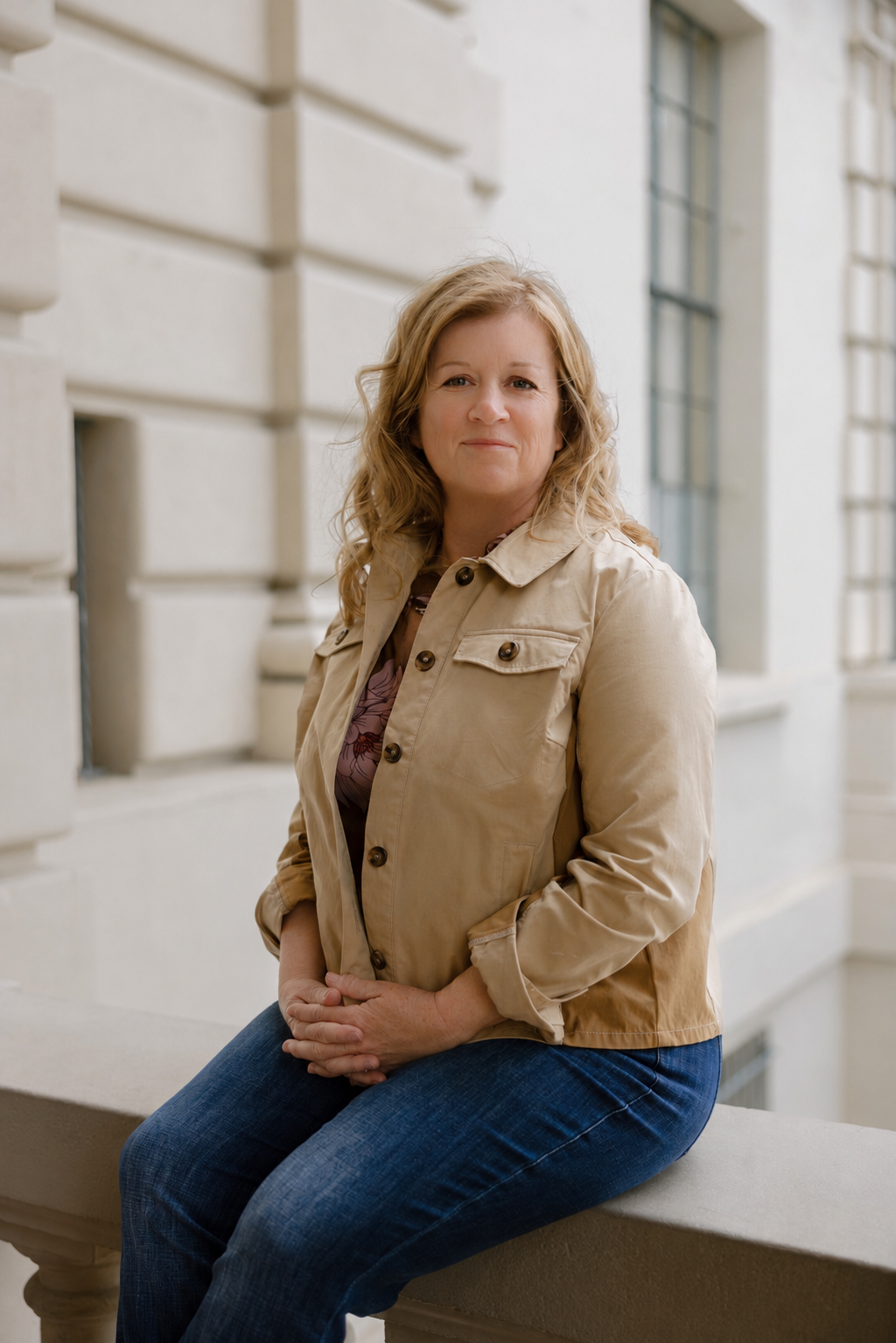A woman with blonde curly hair sitting outdoors on a stone ledge in front of a beige stone building with large windows.
