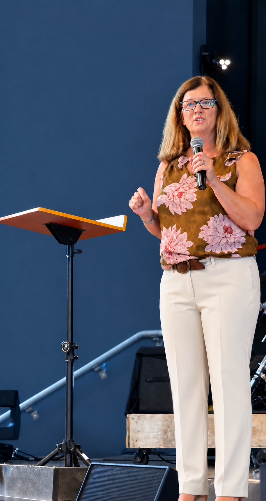A woman wearing glasses and a floral sleeveless top, standing on stage, speaking into a microphone.