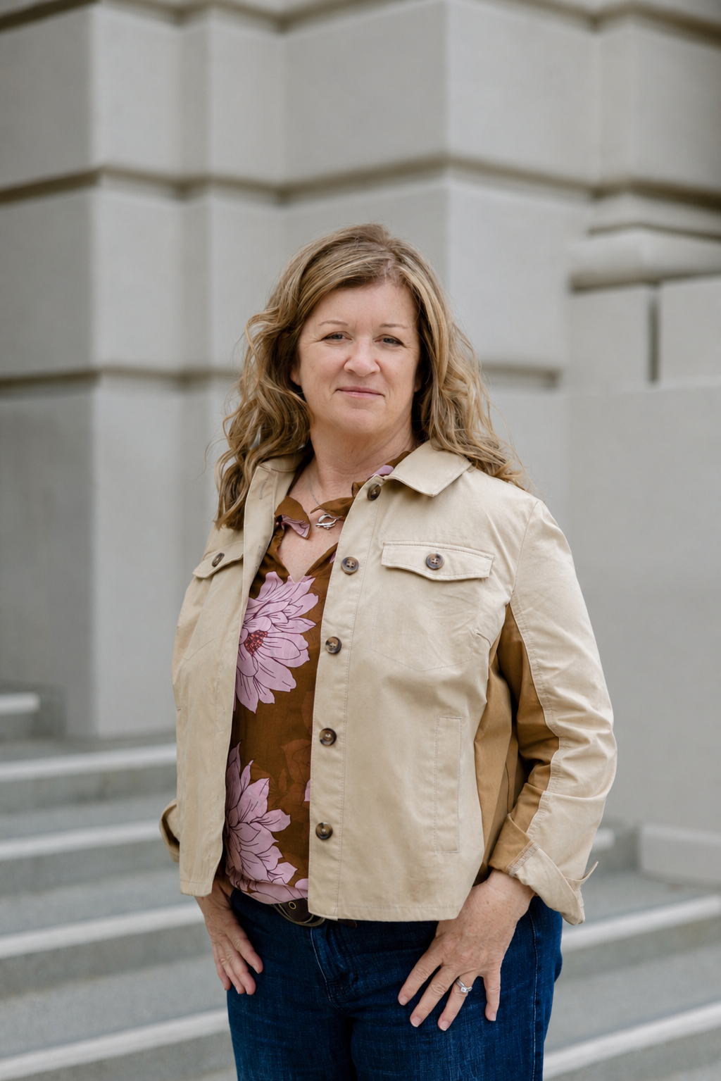 A middle-aged woman with curly blonde hair standing outdoors in front of a modern concrete building, wearing a beige jacket over a pink and brown floral blouse and dark jeans.