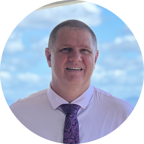 Student Counsellor at Healing Hands Natural Health Centre, a smiling man with short hair, dressed in a light purple shirt and a purple patterned tie, standing outdoors with a blue sky and white clouds in the background.