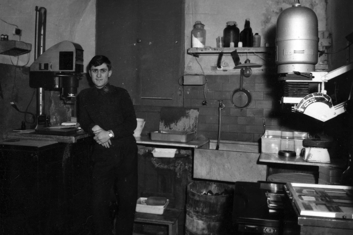 Fotografo in posa in una camera oscura artigianale, anni '60, con ingranditore fotografico e bacinelle di sviluppo, foto in bianco e nero.