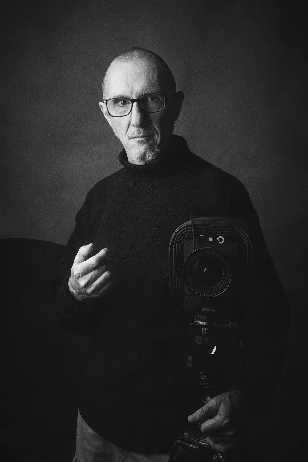 Ritratto bianco e nero in studio di Vieri Bottazzini con maglione nero e occhiali, che tiene uno scatto flessibile davanti a una fotocamera a pellicola in primo piano