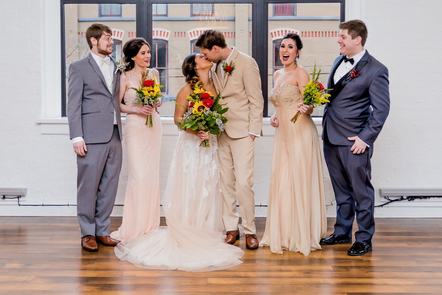 bride and groom kissing in the middle of two bridesmaids in tan dresses and two groomsmen in gray suits.