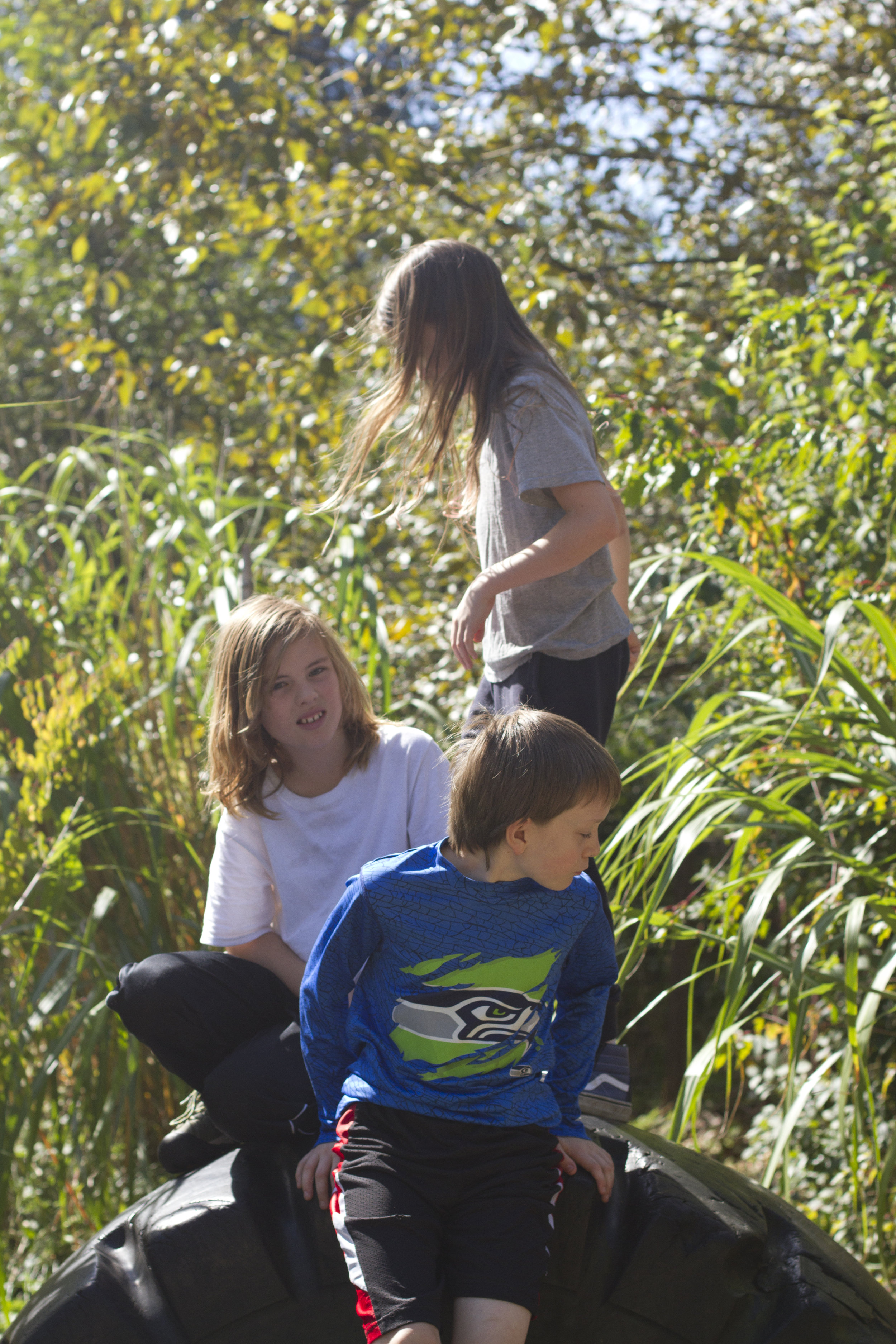 Three students sit or stand on a large tire embedded in the ground.