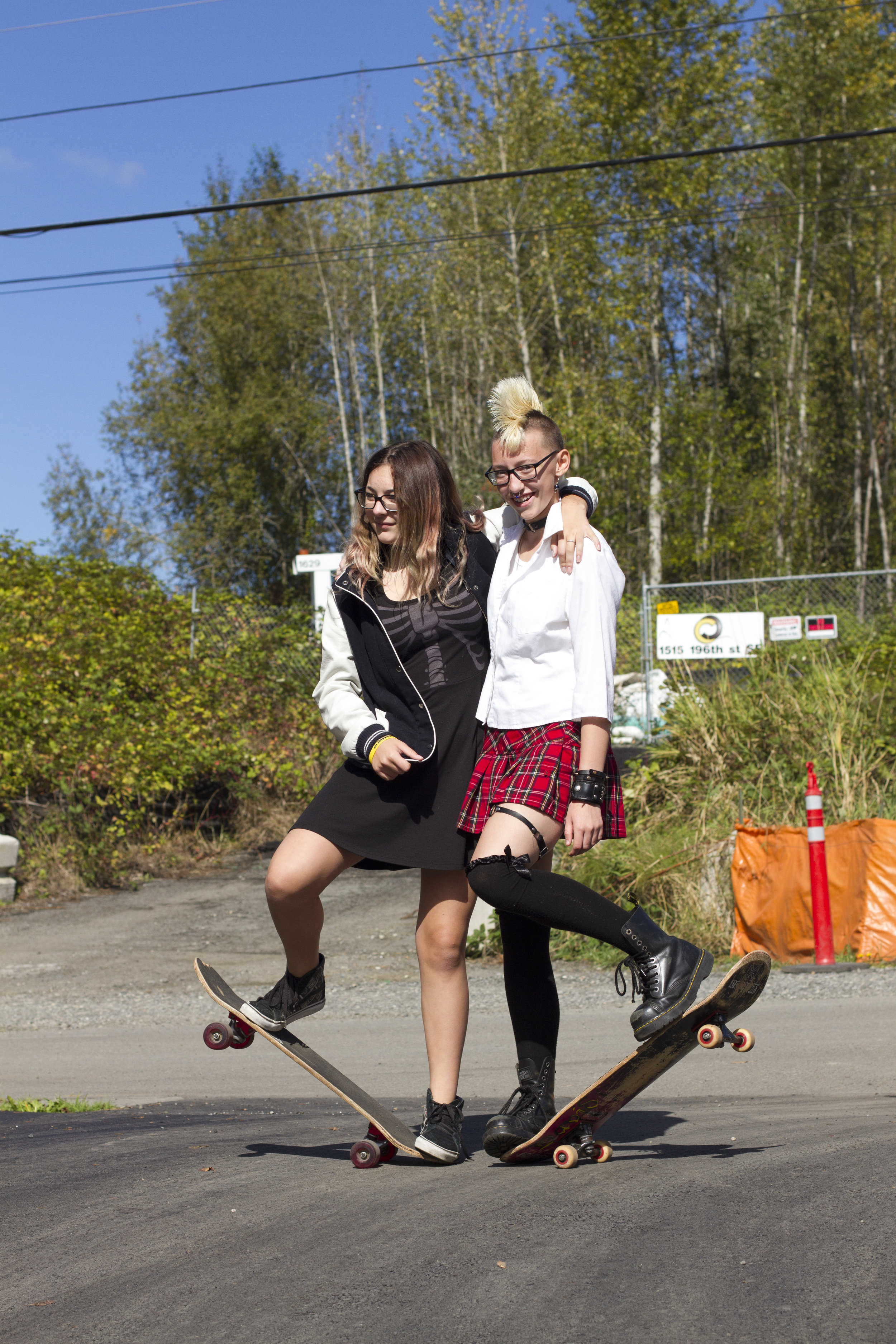 Two students pose with one arm around each other. They each have one foot pushing a skateboard off the ground and the other foot balancing on it.