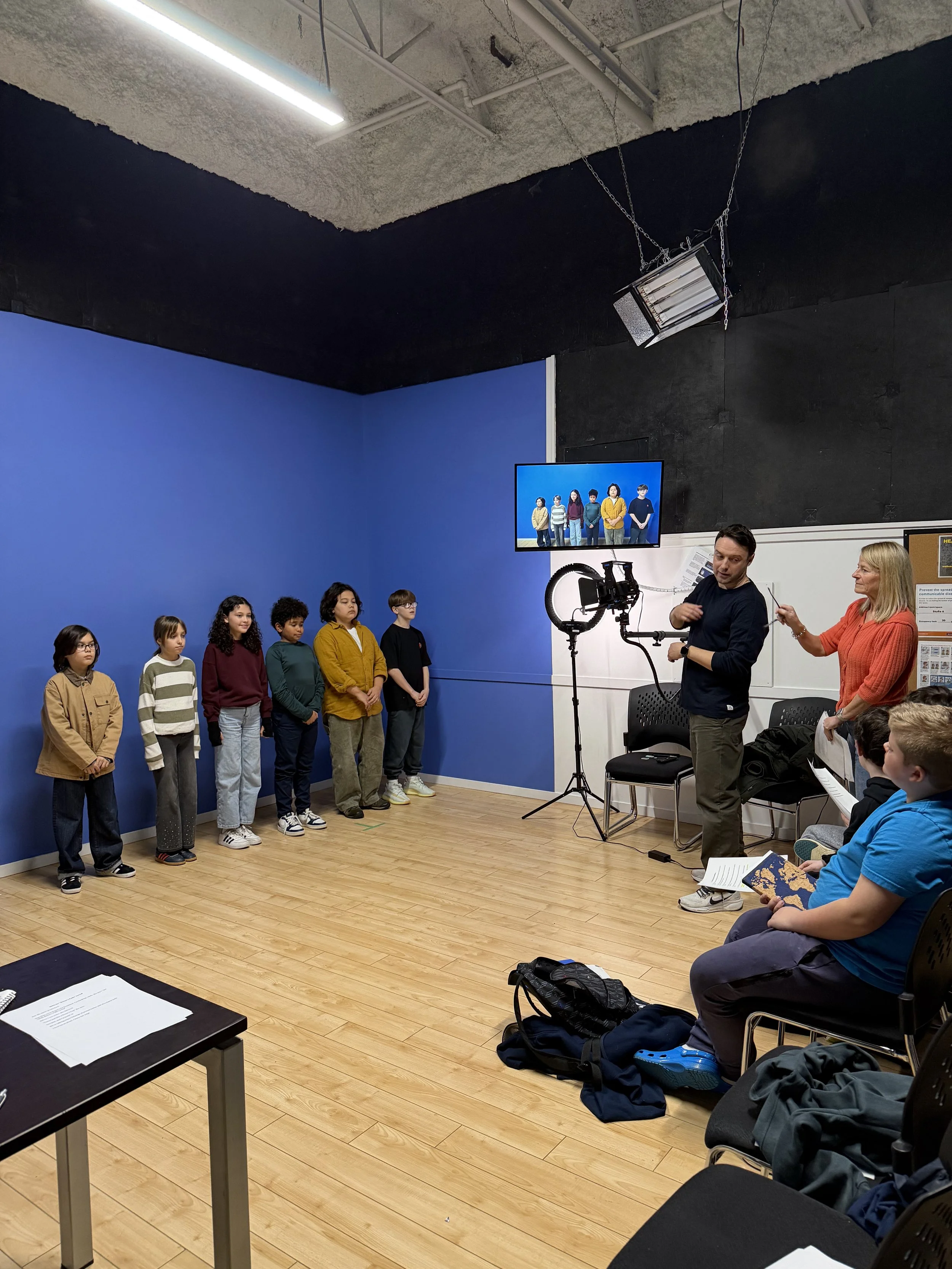 A bunch of child actors standing in front of a blue wall, with Kathleen Mayrs directing