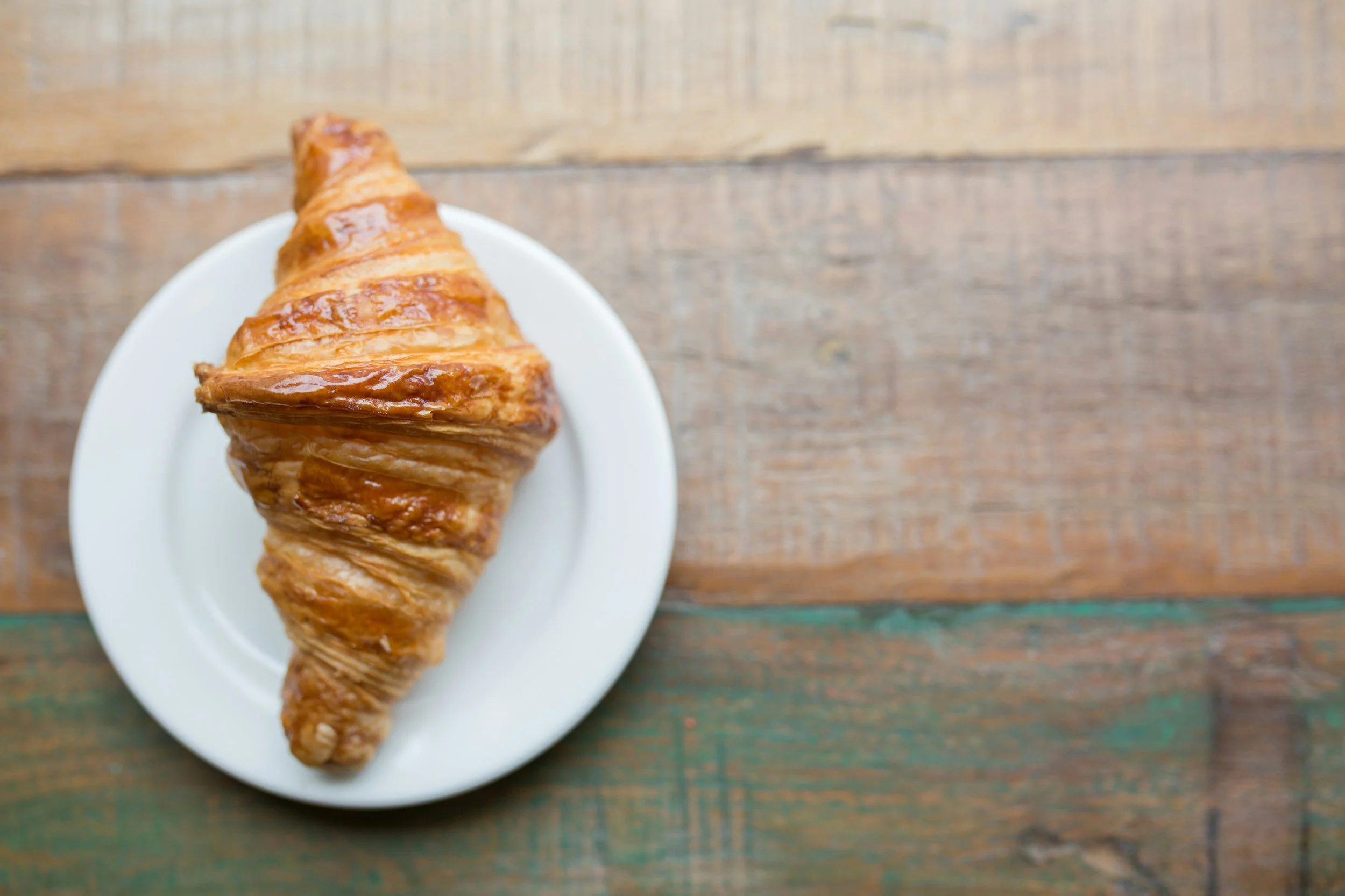 A golden-brown croissant on a white plate on a wooden table.